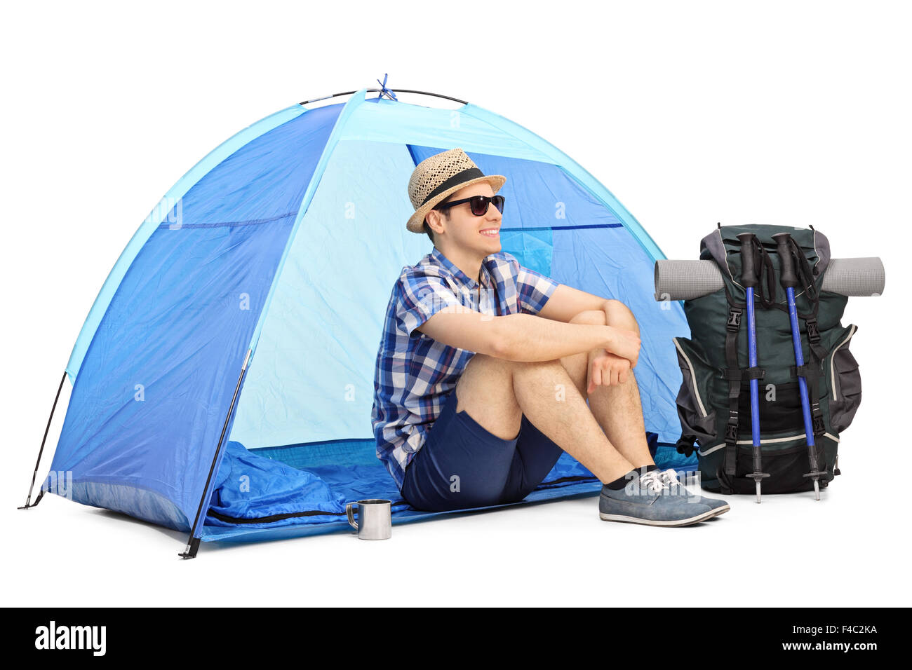 Cheerful male hiker sitting on the floor in front of a blue tent with his backpack and hiking equipment beside him Stock Photo