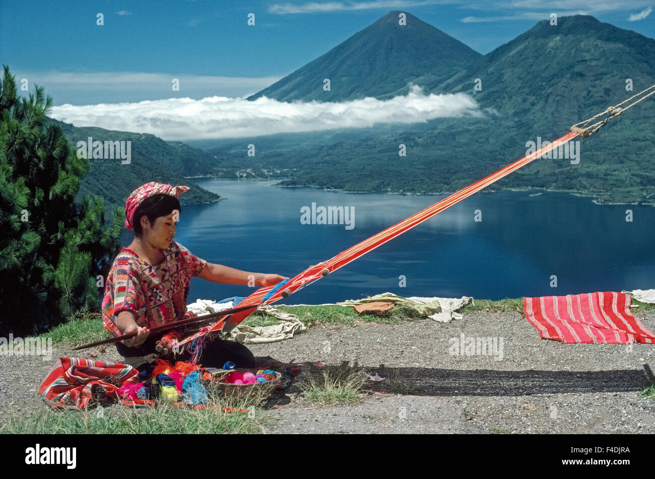 A Mayan girl in native dress weaves at her backstrap loom while overlooking Lake Atitlan in Guatemala. Flanked by volcanoes, Lake Atitlan is the deepest body of water in Central America (1,120 feet/341 meters) and considered one of the most beautiful lakes in the world. Stock Photo