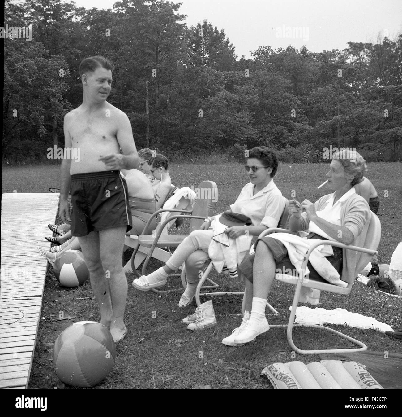 Women sitting on lawn chairs during the summer next to pool with young man talking with them Stock Photo