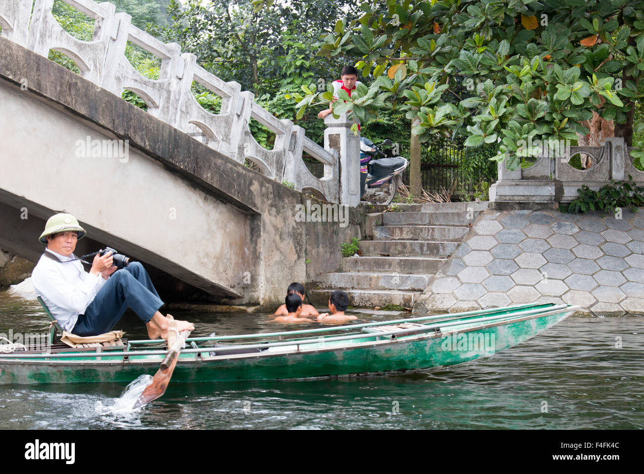 three boys swim bathe in ngo dong river whilst man waits to photograph tourists in the row boats,Ninh binh ,Vietnam Stock Photo