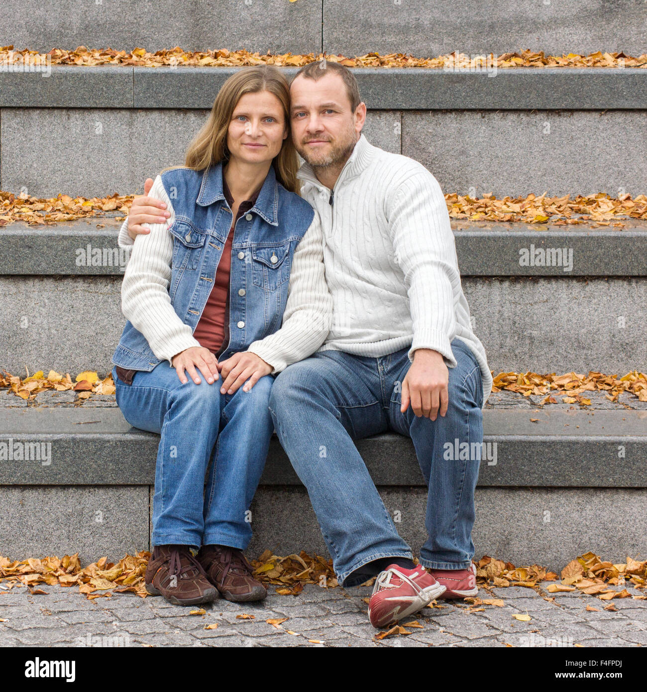 A woman and a man with long hair sitting on gray stone staircase with yellow autumn leaves Stock Photo