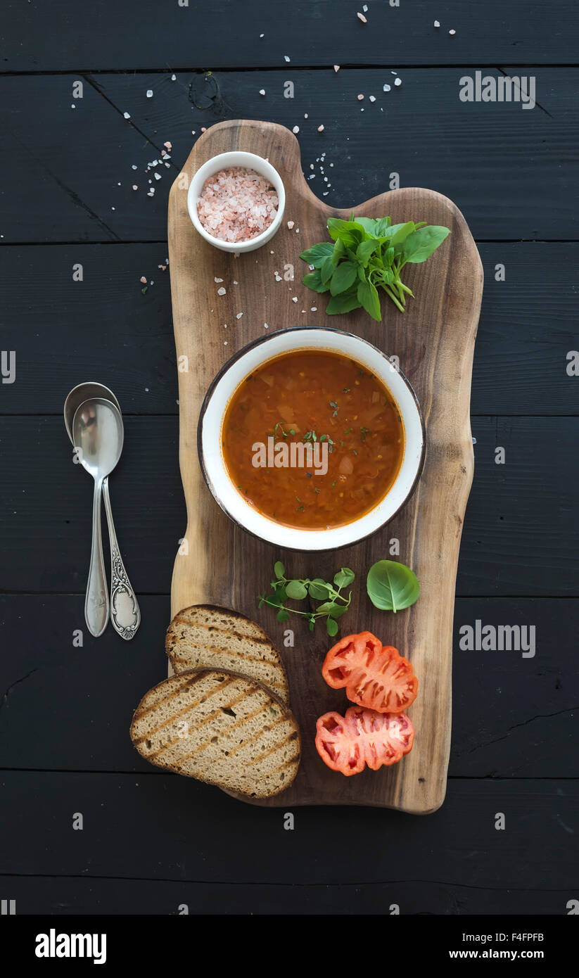 Roasted tomato soup with fresh basil, spices and bread in vintage metal bowl on wooden board over black background, top view Stock Photo
