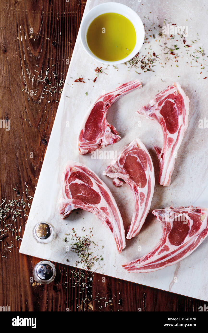 Raw lamb chops with salt, pepper, dry herbs and bowl of olive oil on marble board over wooden table. Top view. Stock Photo