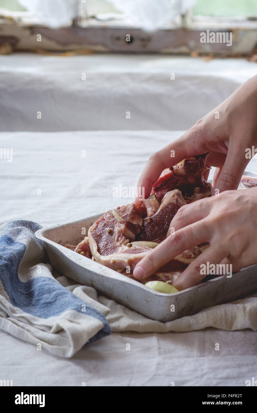 Woman's hand stirring marinated raw lamb chops with onion and pepper in old aluminum baking dish. Over kitchen table with white Stock Photo