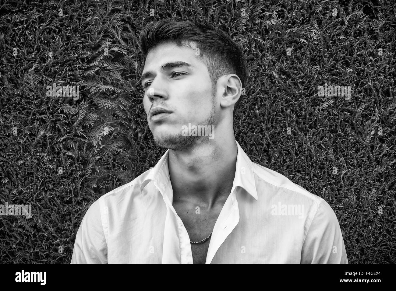 Handsome young man in elegant white shirt standing outdoor in front of green bushy hedge looking away to a side. Black and white Stock Photo