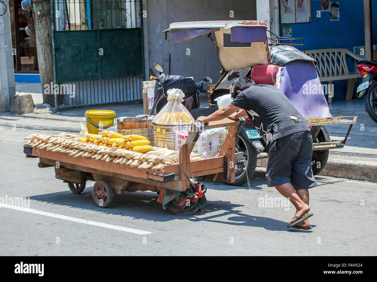 A Filipino man pushes a flat cart along a city road. It's loaded with cooked corn that he sells to people on the street. Stock Photo