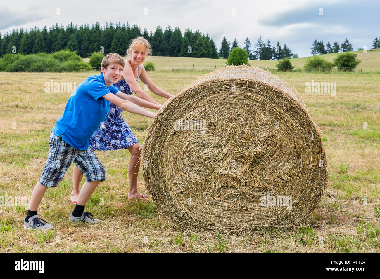 Woman and teenage boy  rolling hay bale on grass in rural area Stock Photo