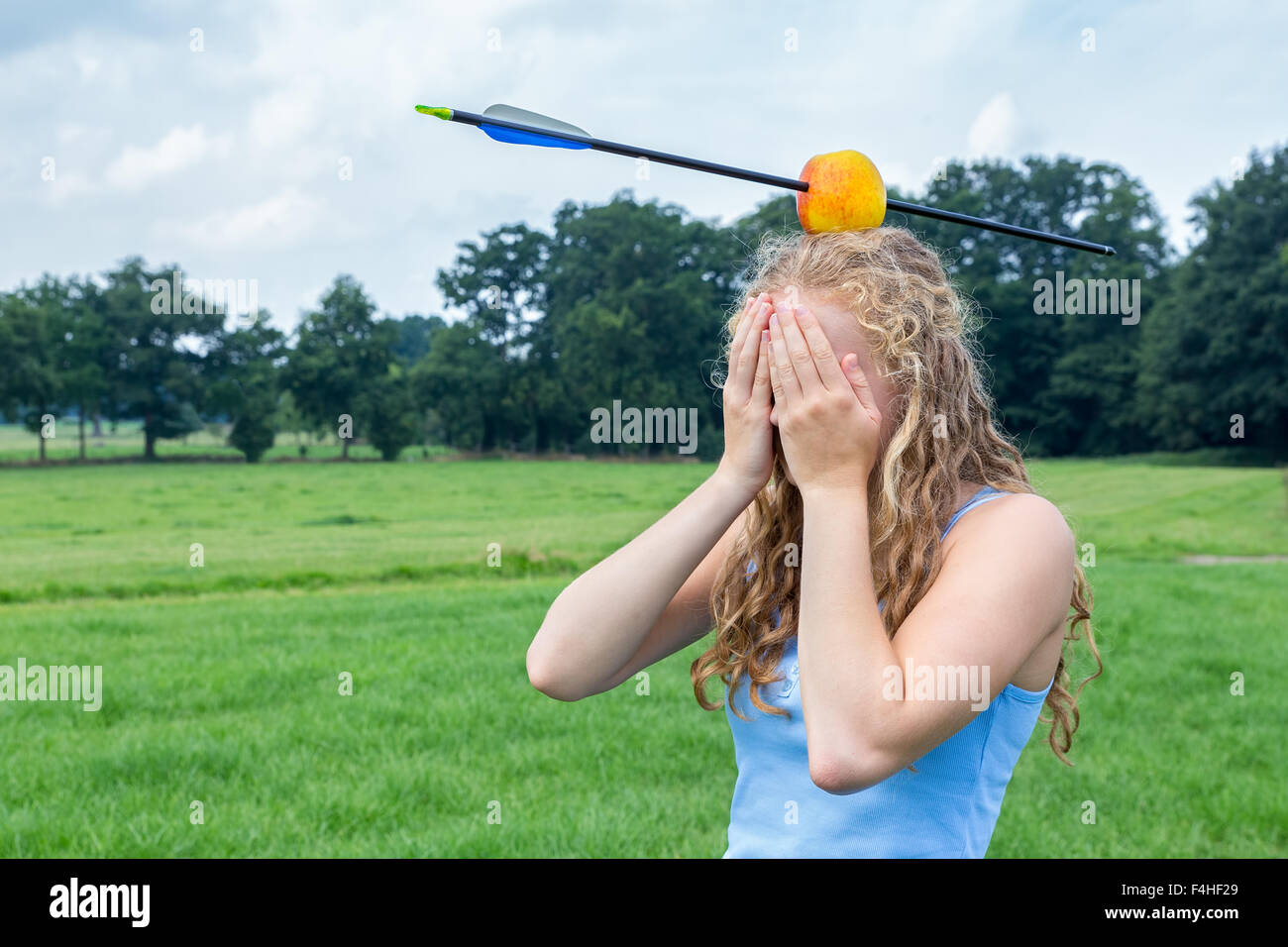 Teenage girl covering her face feeling fearful with apple and arrow on head in green meadow outdoors Stock Photo