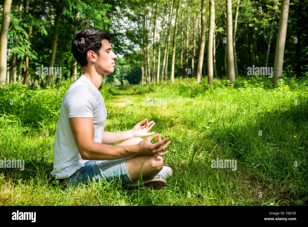 Profile of handsome Young Man During Meditation or Doing an Outdoor Yoga Exercise Sitting Cross Legged on Grassy Ground Alone in Stock Photo