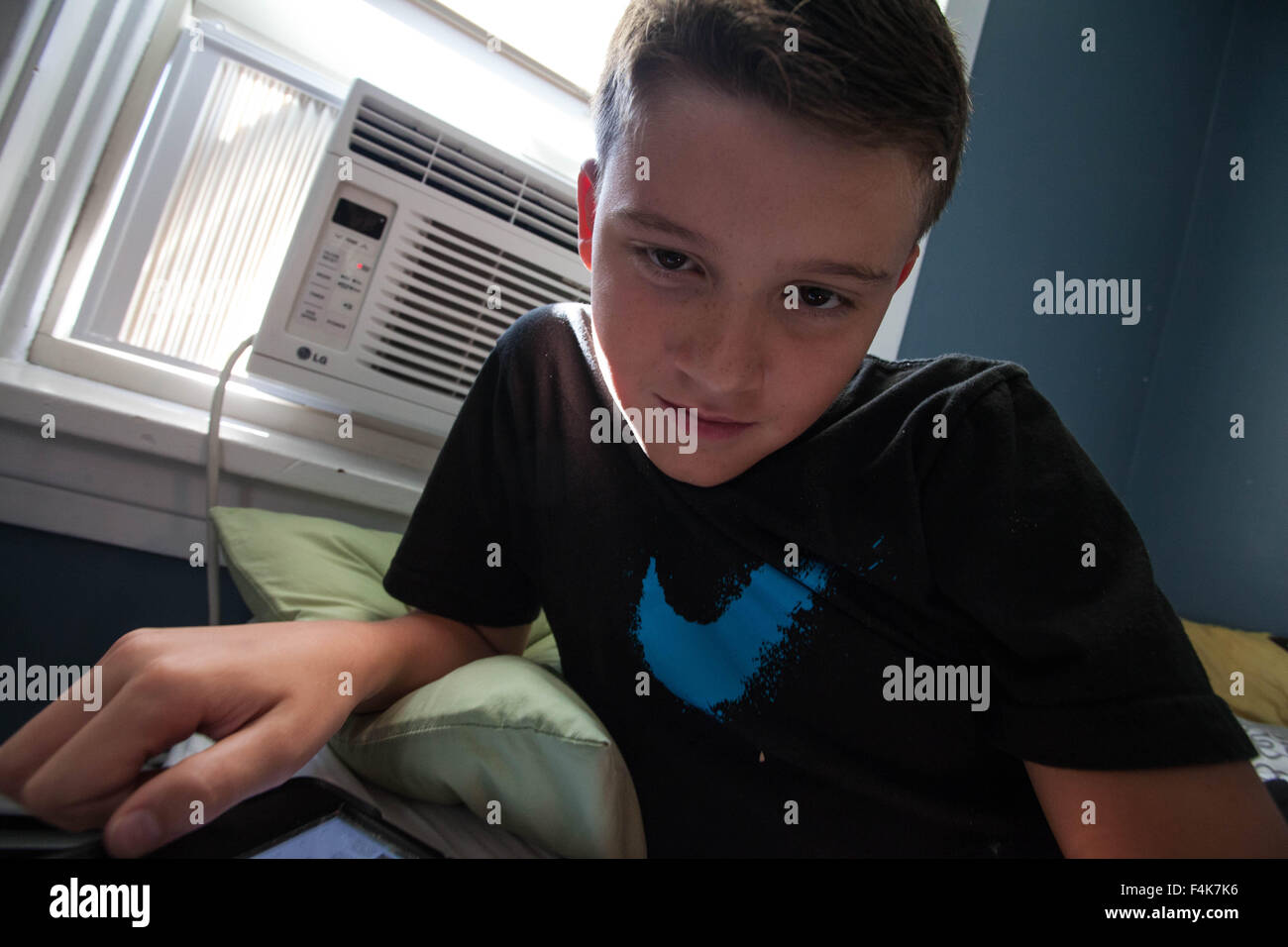 A young boy looks at his tablet alone in his bedroom on a sunny day Stock Photo
