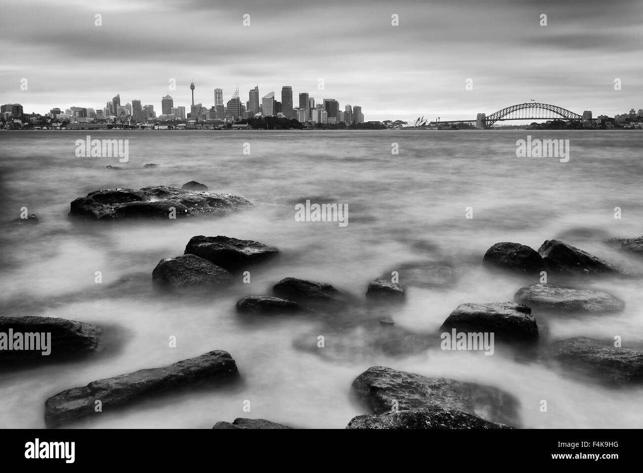 Distant Sydney city CBD landmarks with Harbour Bridge across blurred harbour waters at sunset with beach rocks in foreground Stock Photo