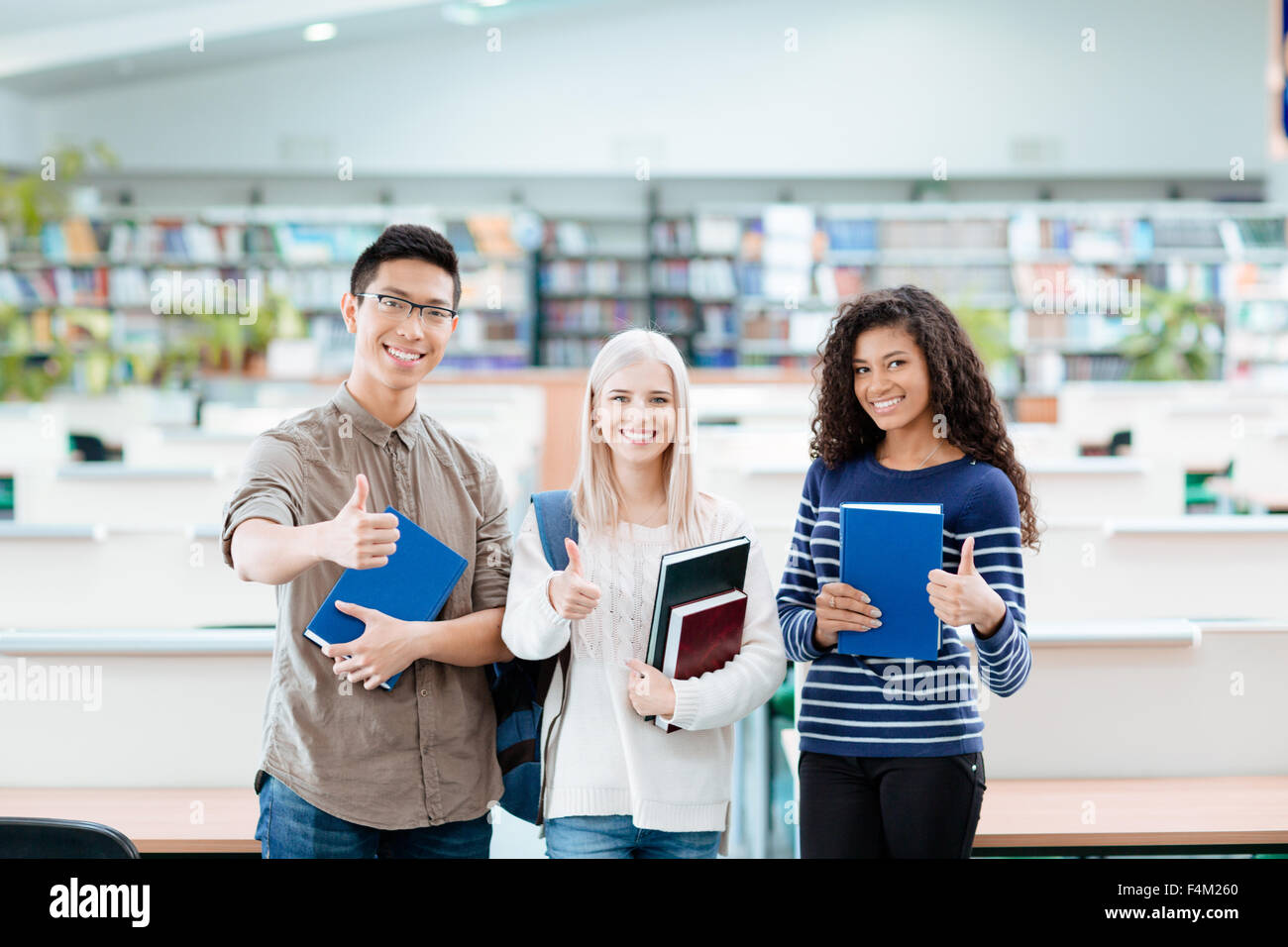 Portrait of a happy multi ethnic students standing in university library and showing thumbs up Stock Photo