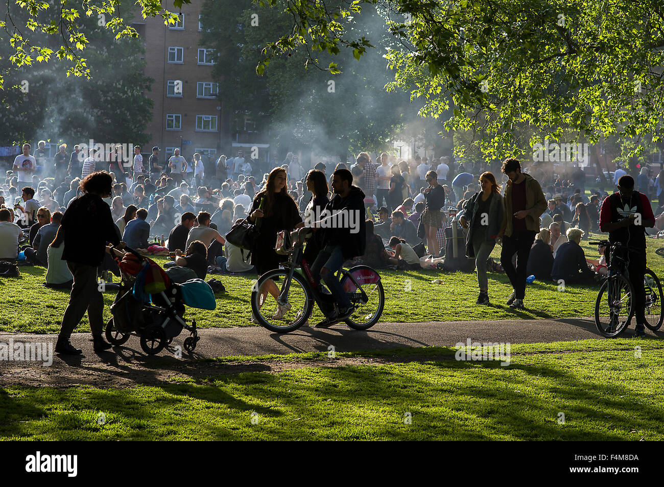 Large group of people in London Fields park on a sunny day, sitting on the grass, barbecuing, clouds of smoke rising Stock Photo