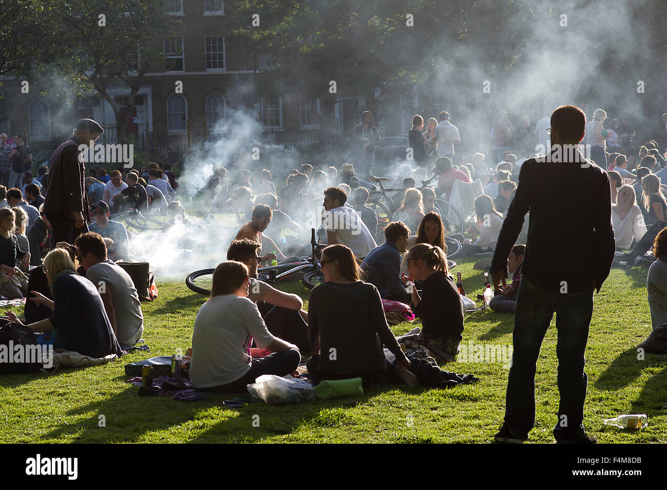 Large group of people in London Fields park on a sunny day, sitting on the grass, barbecuing, clouds of smoke rising Stock Photo