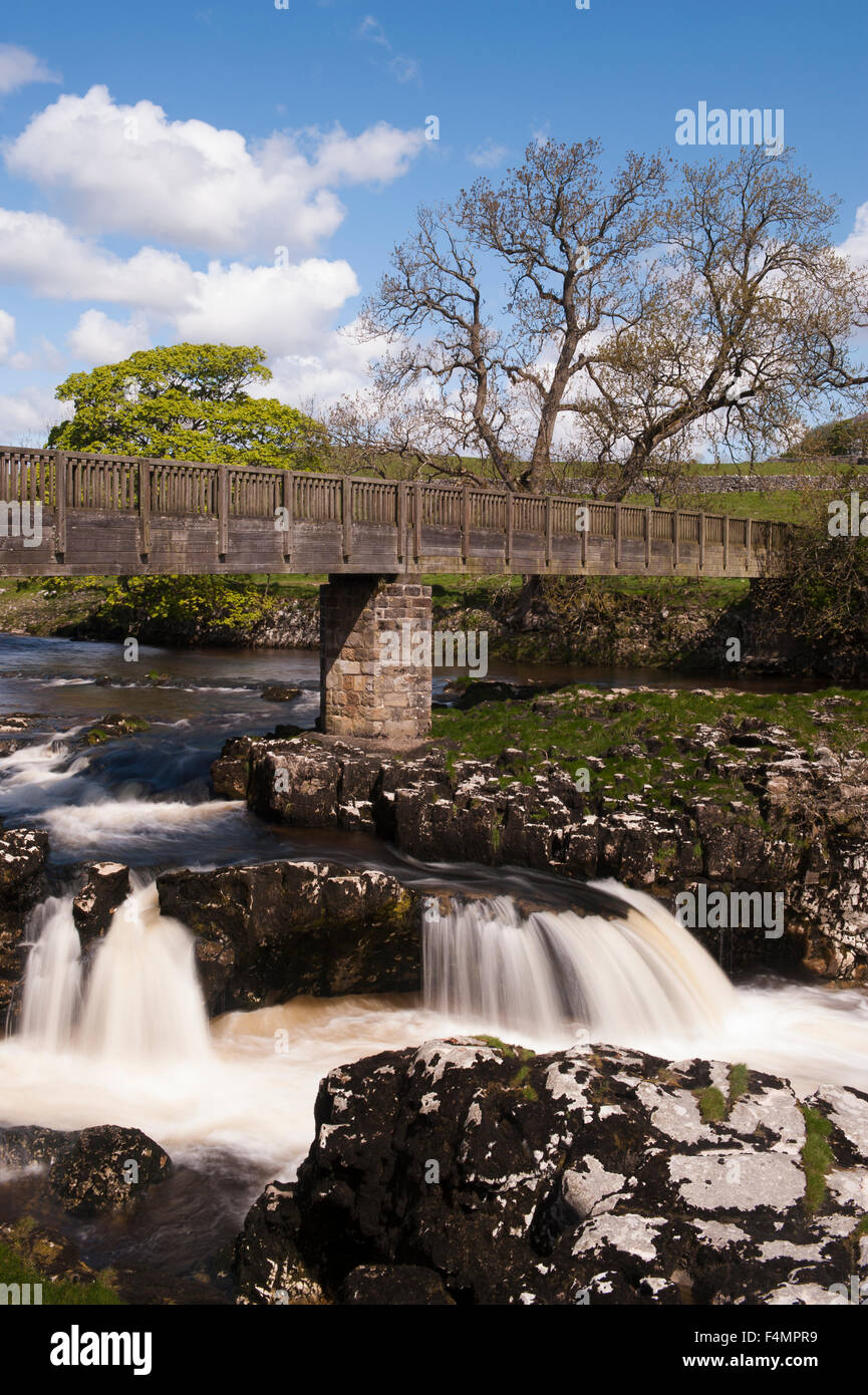 River Wharfe flowing (slow shutter speed view) under footbridge & blue sky - sunny scenic Linton Falls waterfall, Grassington, Yorkshire, England, UK. Stock Photo