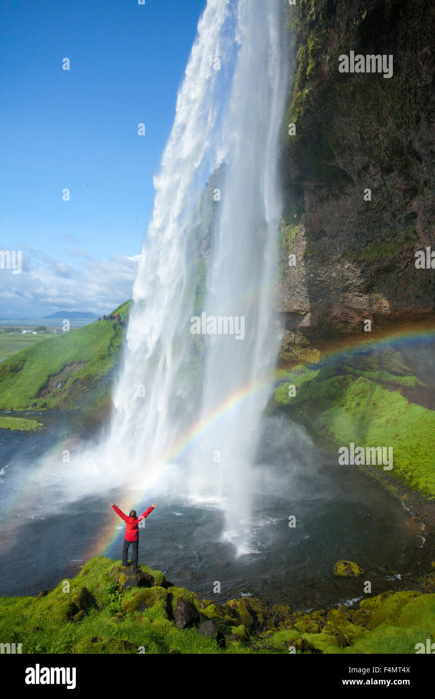 Person and rainbow beneath 60m-high Seljalandsfoss waterfall, Sudhurland, Iceland. Stock Photo