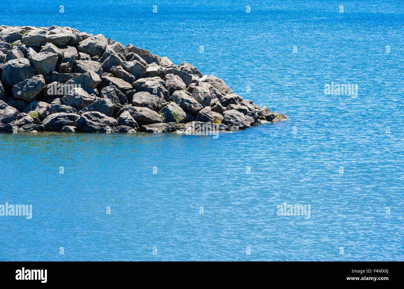 Edge of rocky breakwall descending into shallow cyan water with ripples in light wind. Stock Photo