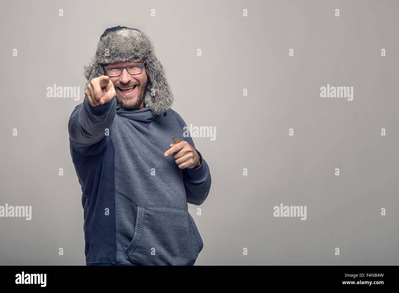 Laughing jovial man wearing glasses and a furry winter hat standing pointing at the camera with a playful expression, over grey Stock Photo