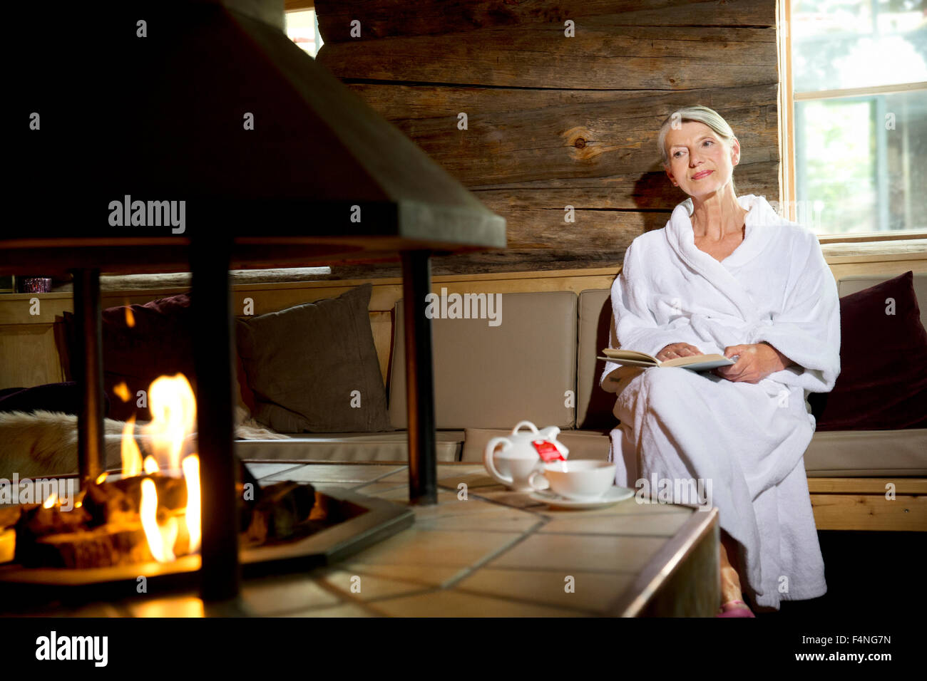 Senior woman sitting on bench in bathrobe at the fireplace Stock Photo