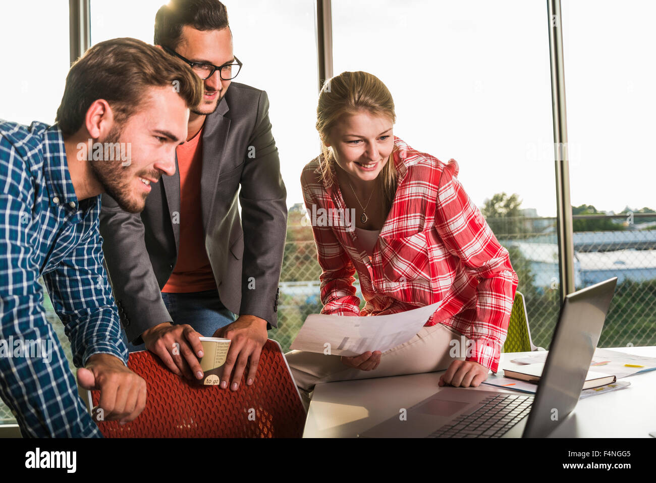 Three smiling young people in conference room looking at laptop Stock Photo