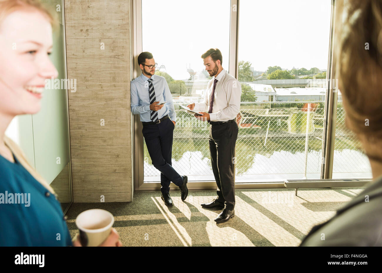 Two young businessmen talking at the window in office with colleagues ...