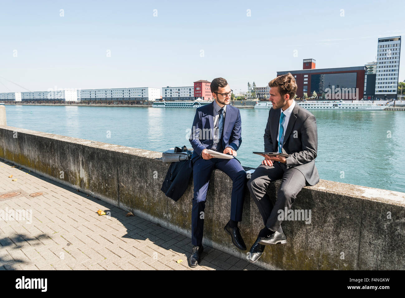 Two young businessmen sitting on wall by river, working Stock Photo