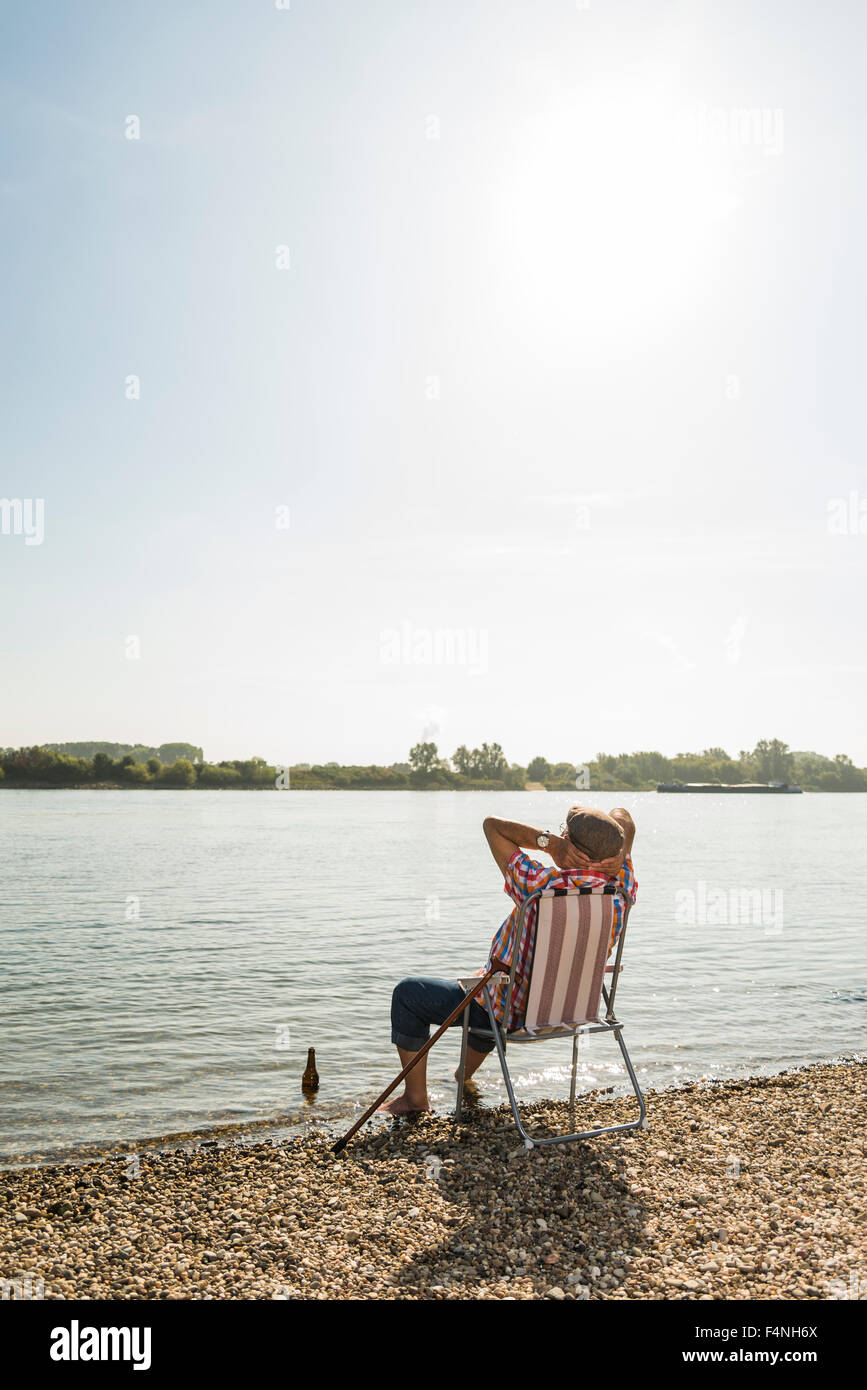 Germany, Ludwigshafen, back view of senior man rrlaxing on folding chair at riverside Stock Photo