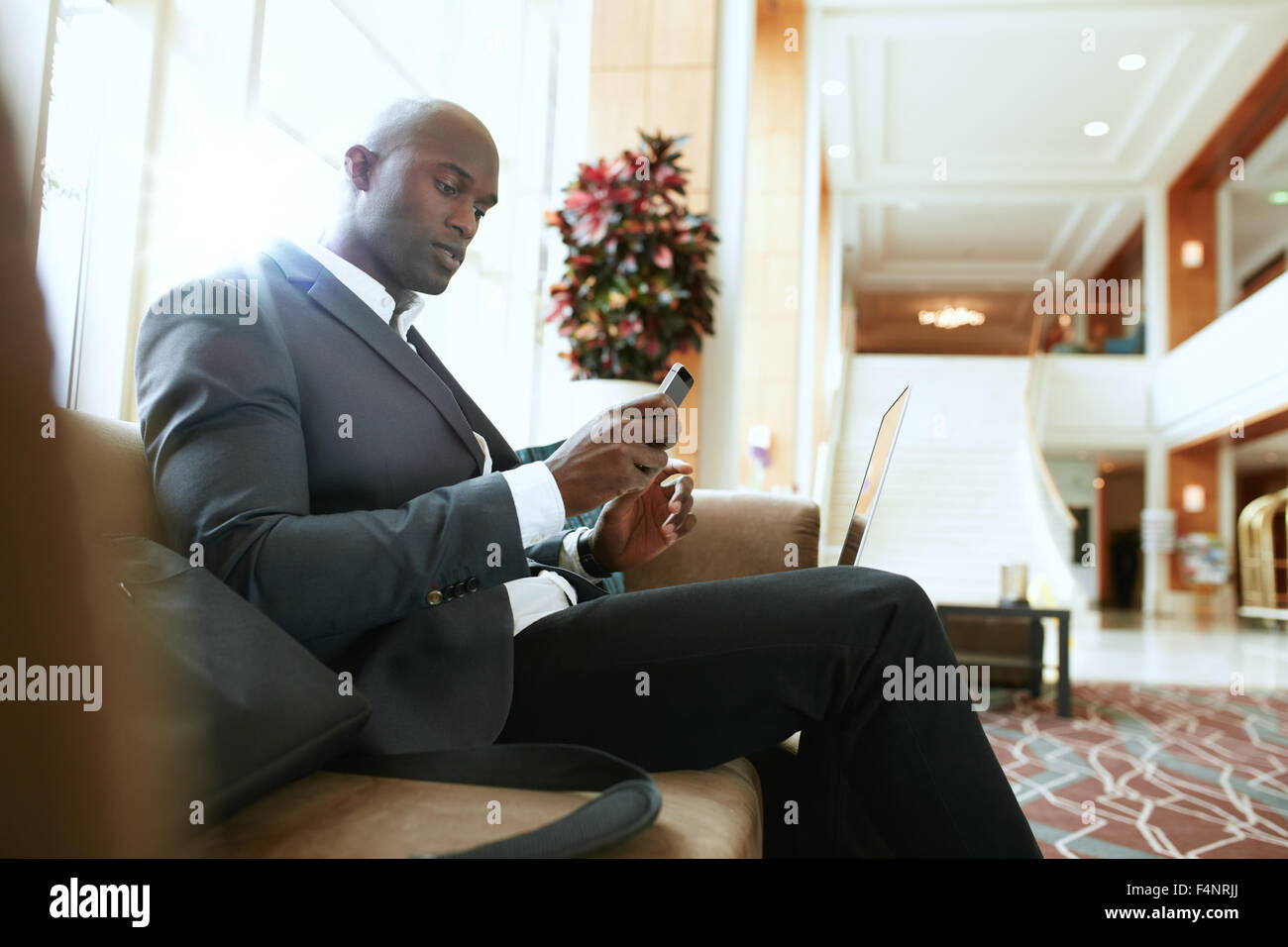 Male executive sitting on sofa looking at his mobile phone. African businessman waiting in hotel lobby. Stock Photo