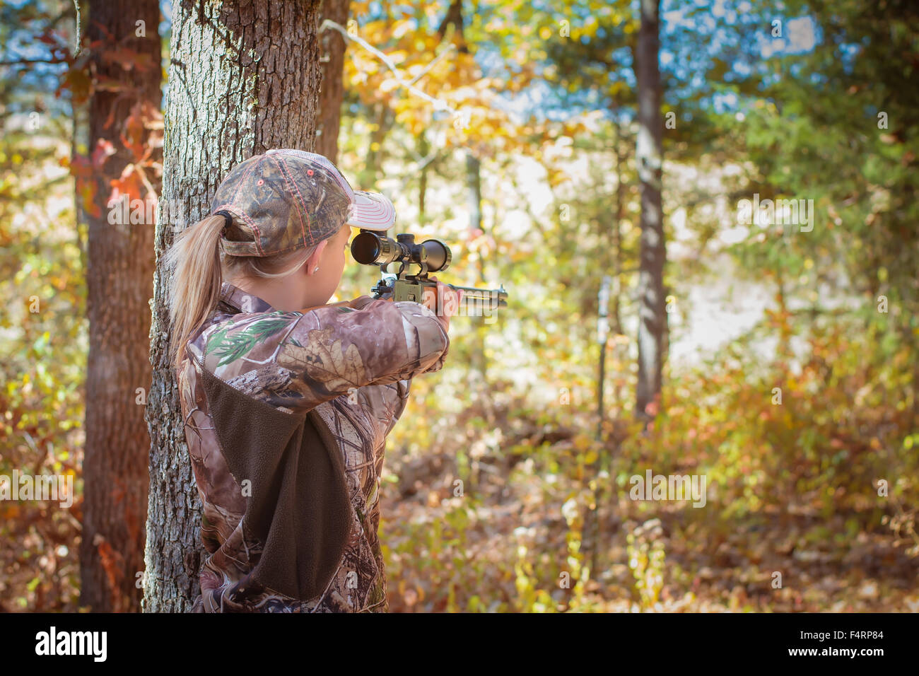 Young woman aiming a rifle in a wooded area wearing camouflage. Stock Photo