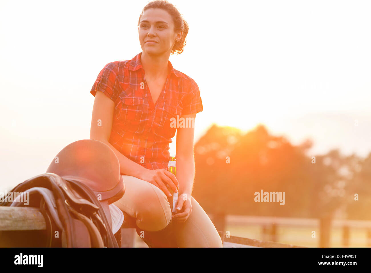 Smiling woman drinking beer next to saddle on rural fence Stock Photo