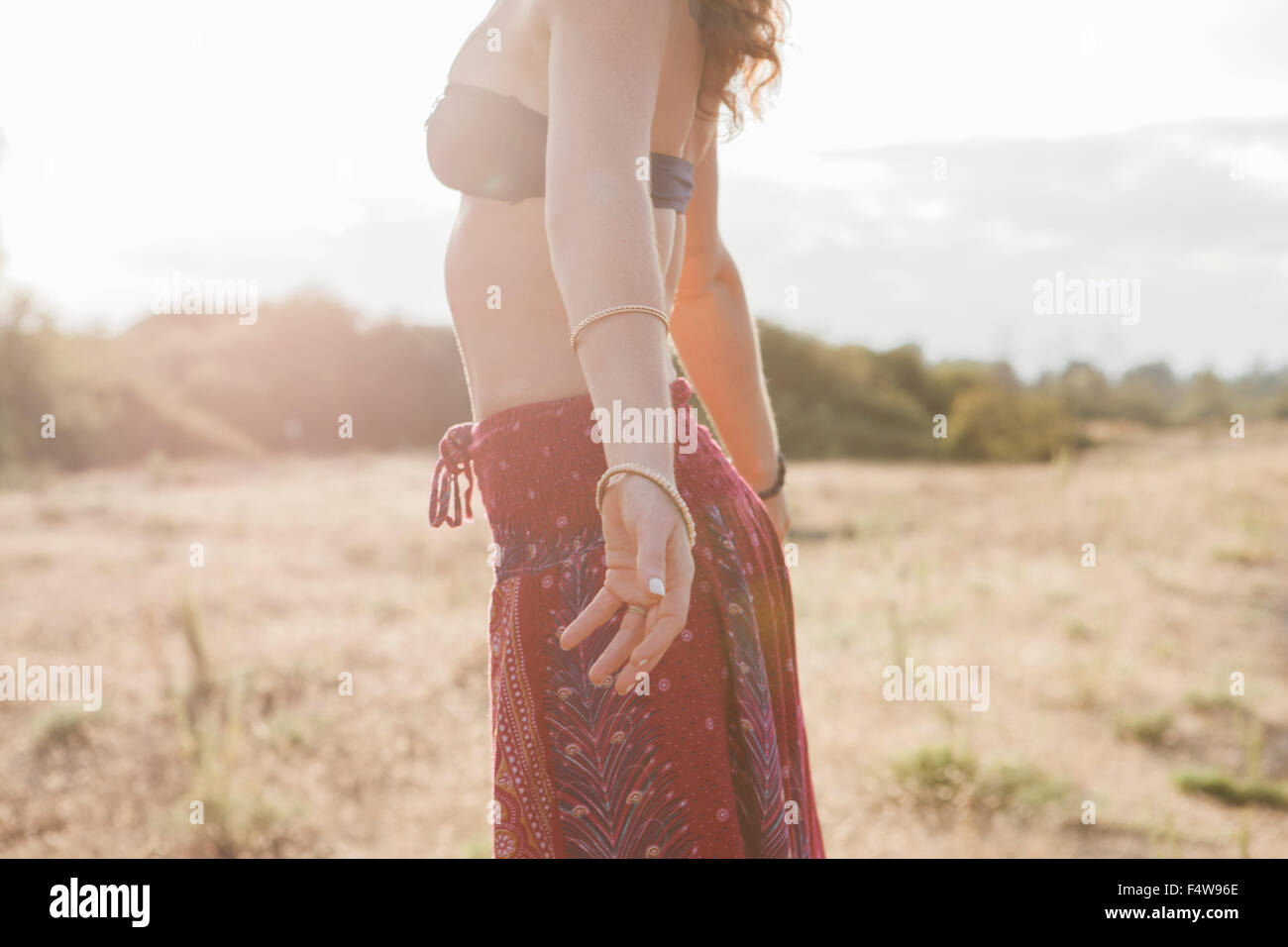 Boho woman in bikini top and skirt with arms outstretched in sunny rural field Stock Photo
