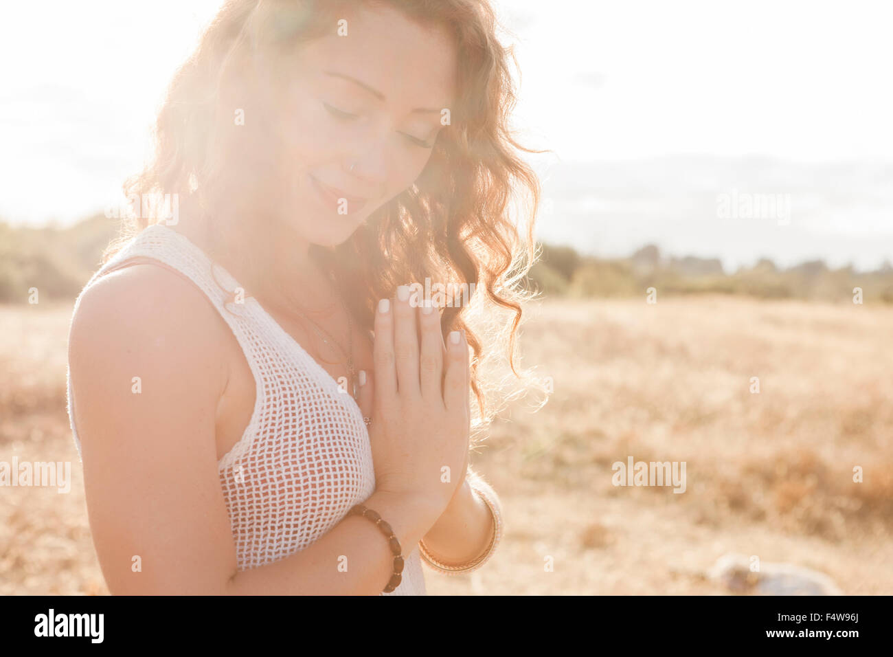 Serene boho woman with hands at heart center in sunny rural field Stock Photo