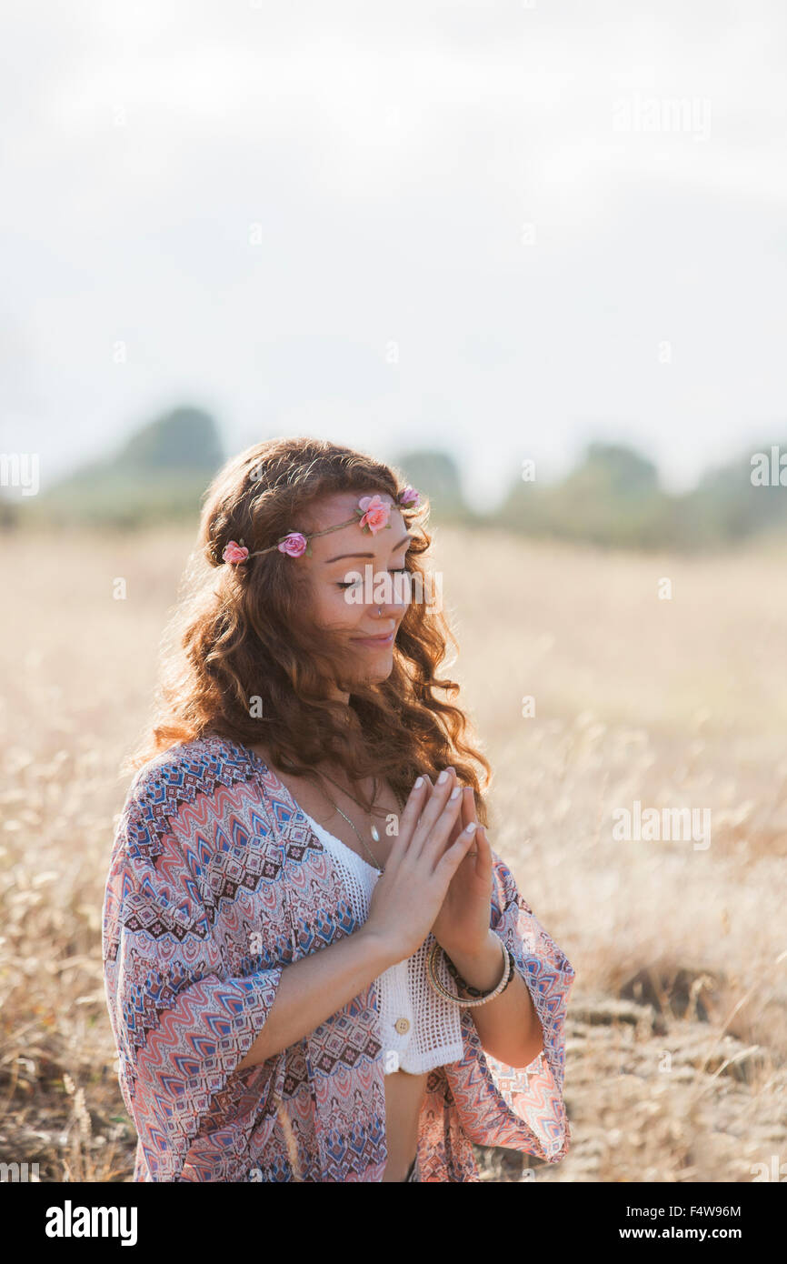 Serene boho woman meditating with hands at heart center in sunny rural field Stock Photo