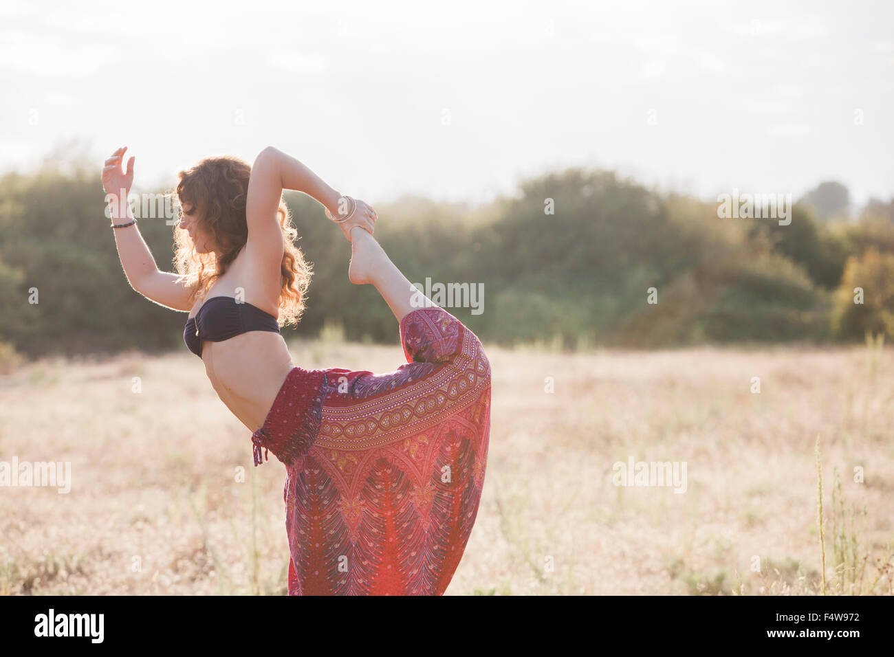 Boho woman in king dancer yoga pose in sunny rural field Stock Photo