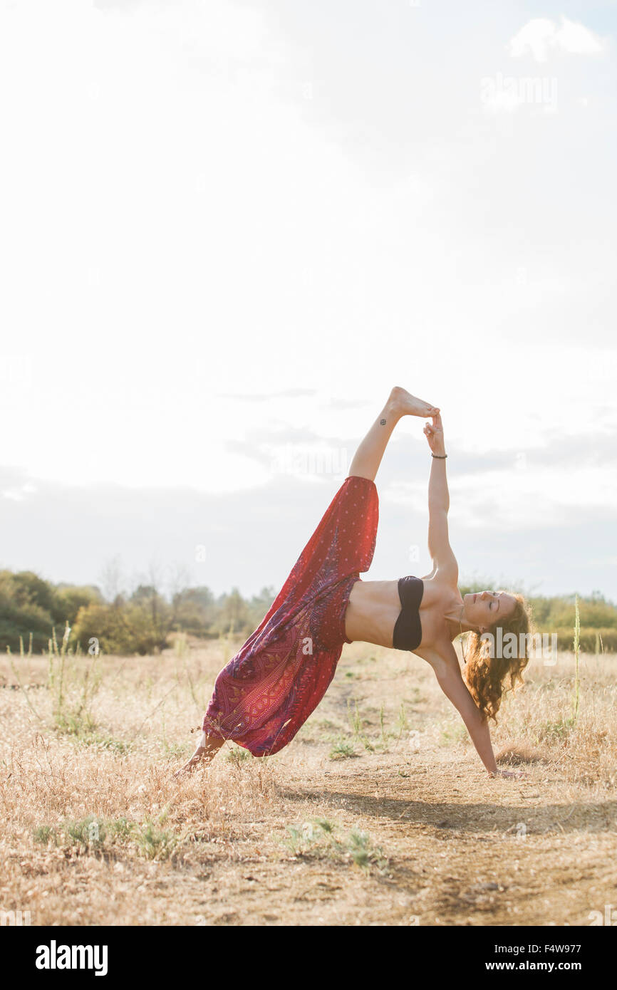 Boho woman in extended side plank yoga pose in sunny rural field Stock Photo