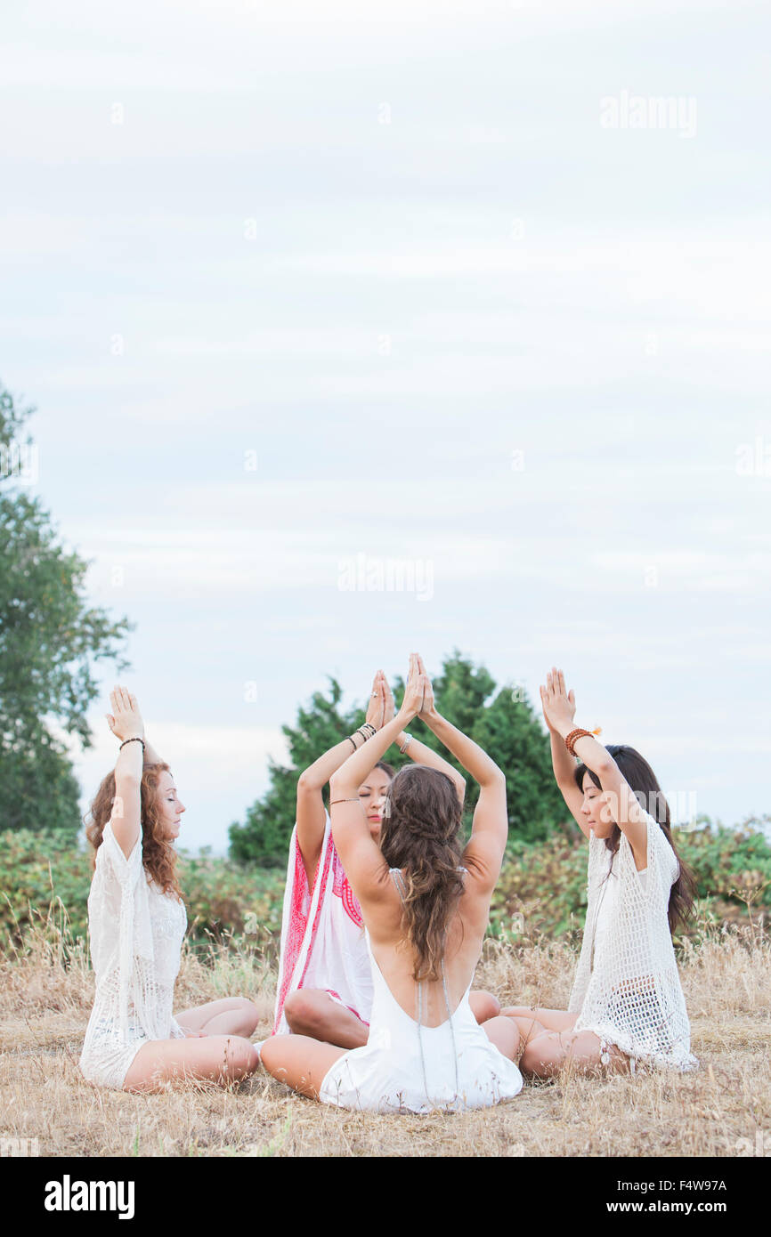 Boho women meditating with hands clasped overhead in circle in rural field Stock Photo
