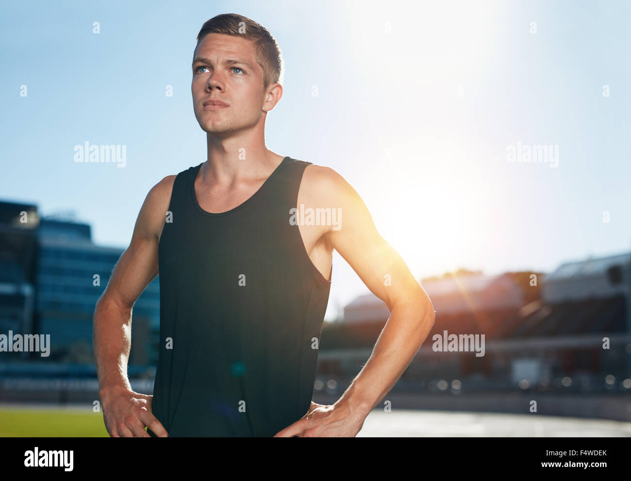 Young male runner standing with his hands on hips looking away. Determined athlete on race track in athletics stadium with sun f Stock Photo