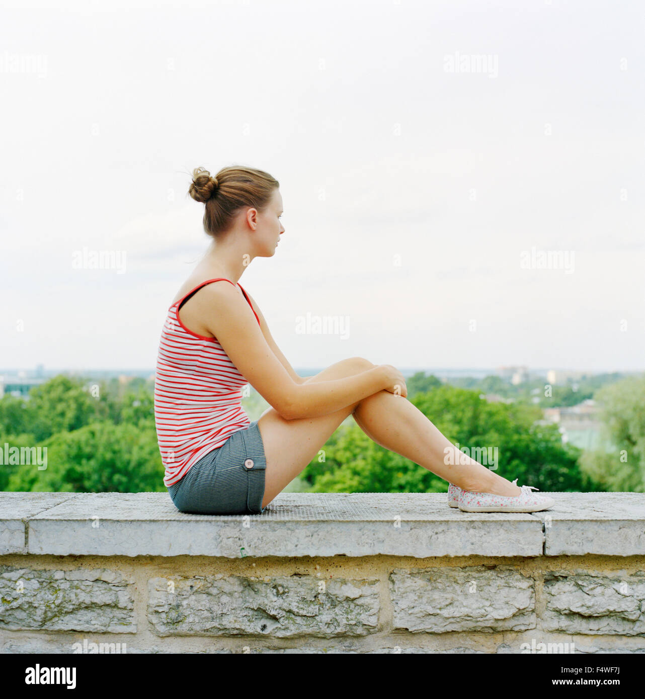 Side view of young woman sitting on surrounding wall and looking at view Stock Photo