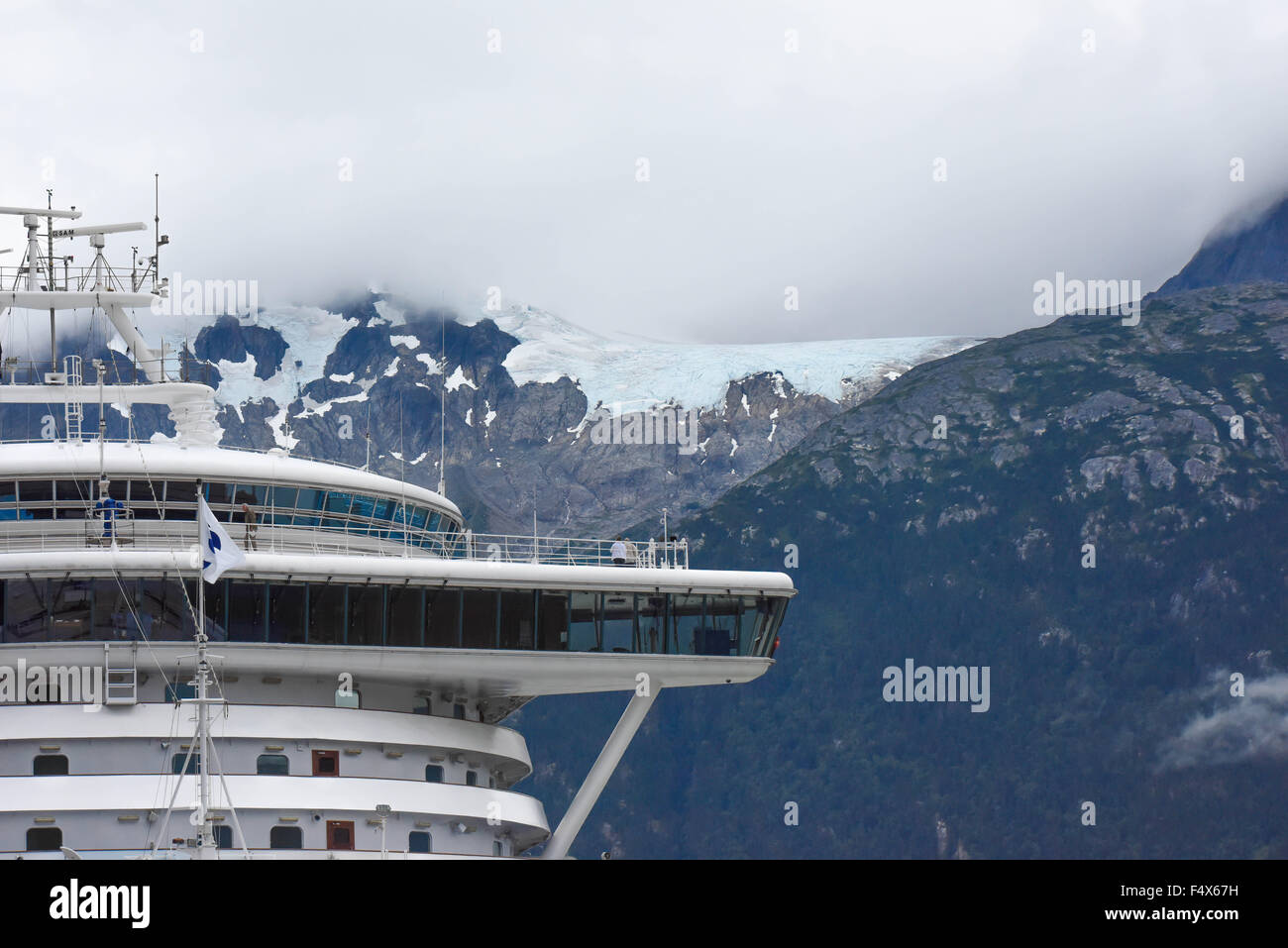 A cruise ship is docked under a hanging glacier and clouds on the mountain tops near port of Skagway Alaska | Lynn Canal fjord cruise - inside passage Stock Photo