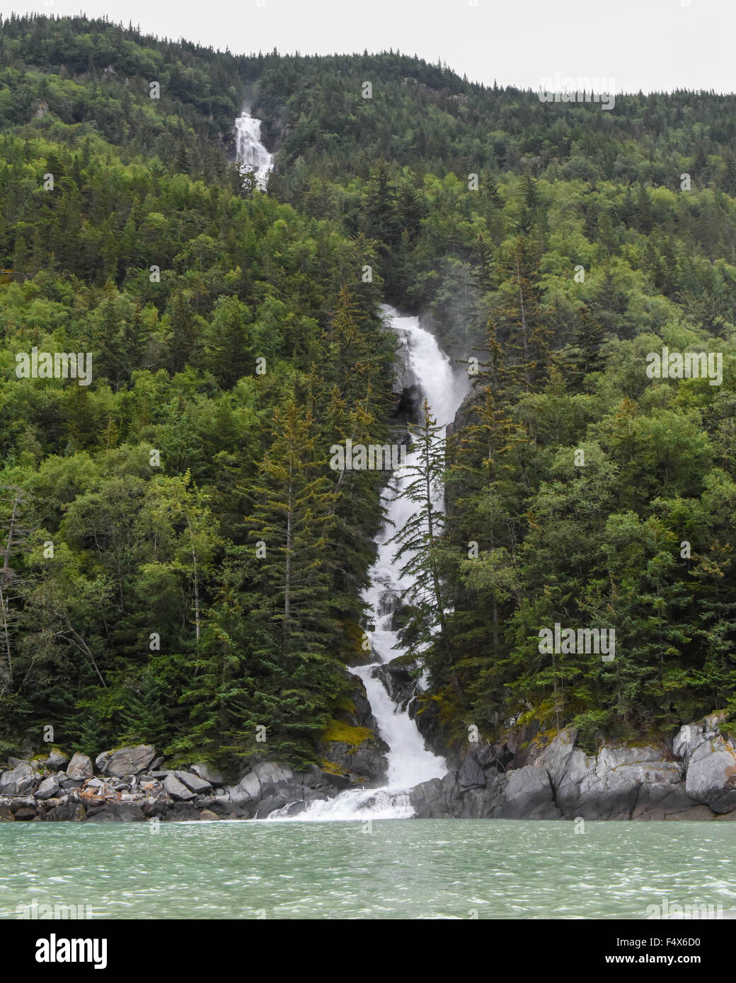 A scenic mountainside waterfall crashes through a forest of pine trees near Skagway Alaska | Lynn Canal fjord cruise - inside passage Stock Photo