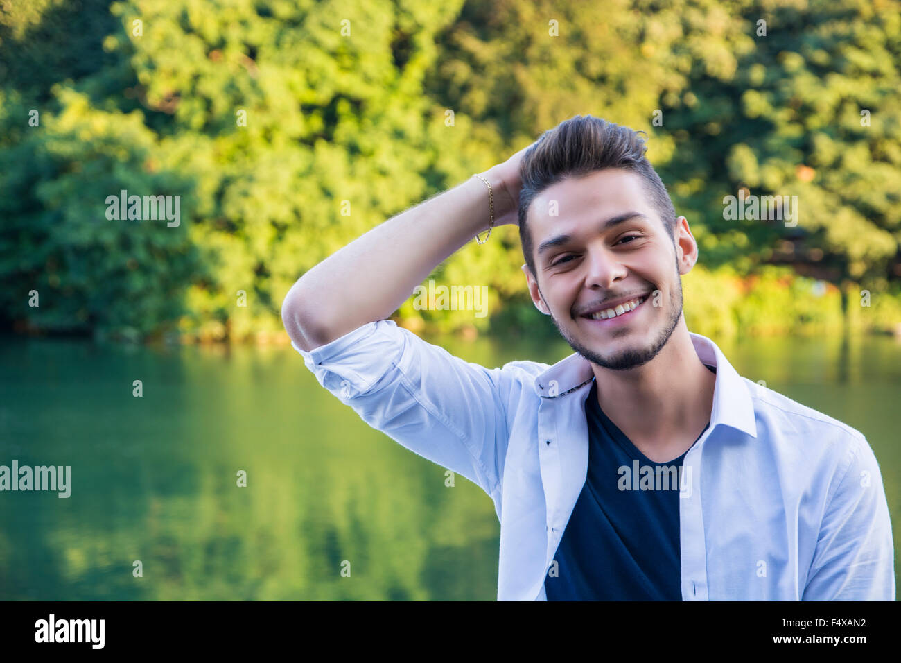 Portrait of contemplative light brown haired young man wearing white shirt beside picturesque river or lake, smiling to camera Stock Photo