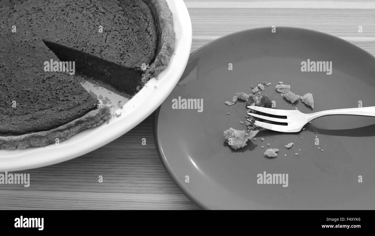 Plate with crumbs and fork next to a sliced pumpkin pie on a wooden table - monochrome processing Stock Photo