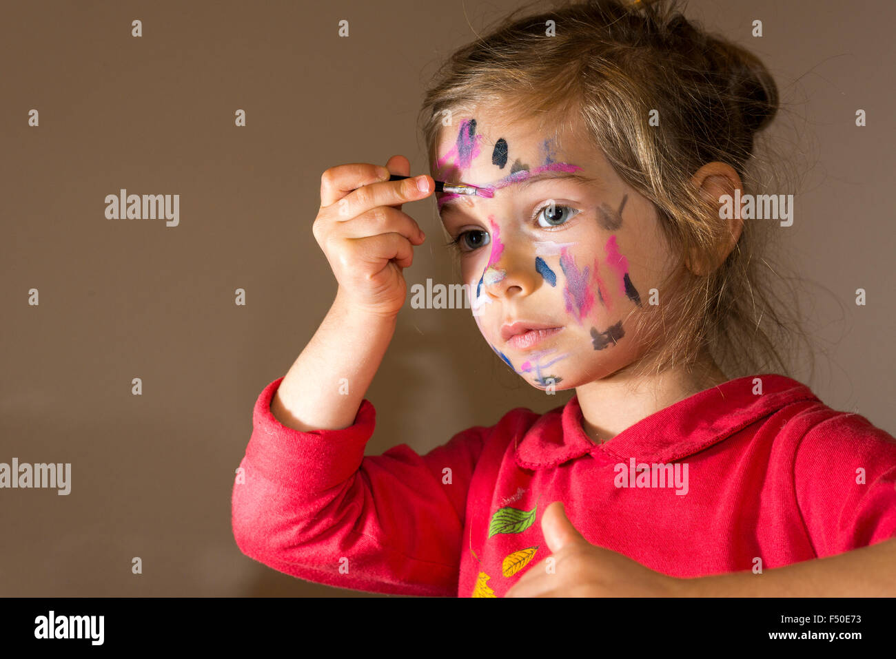 A little girl, wearing a red sweater, is painting her face with colorful watercolor Stock Photo