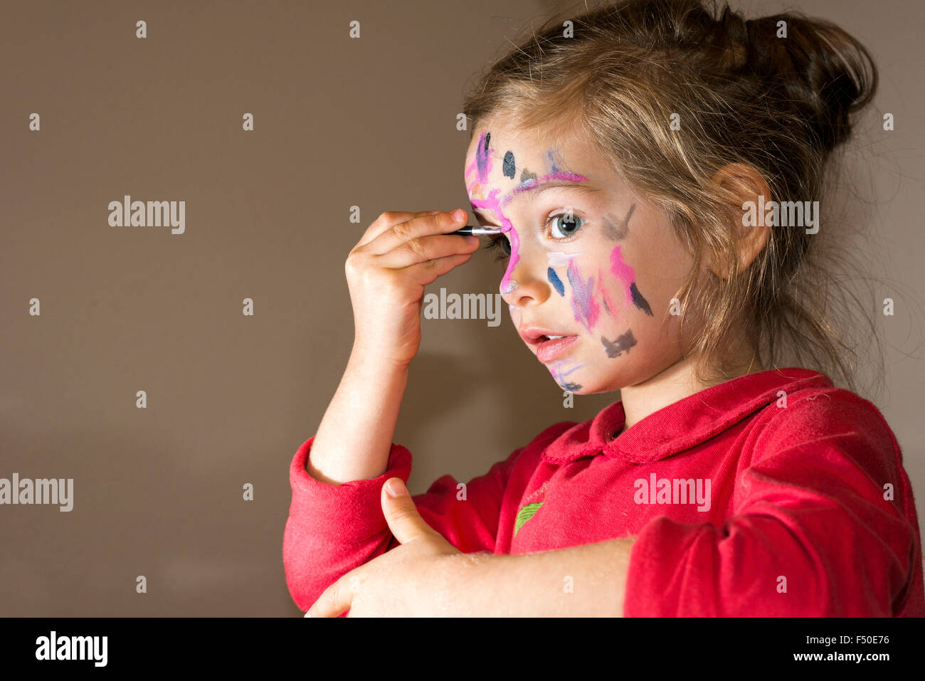 A little girl, wearing a red sweater, is painting her face with colorful watercolor Stock Photo