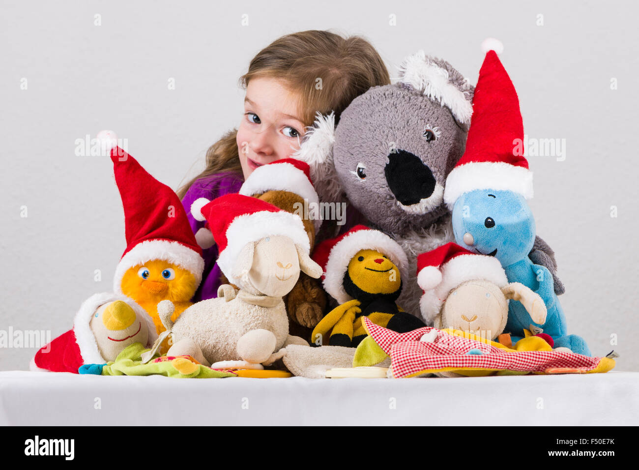 A three year old girl is presenting her favoured colorful cuddly toys, wearing Santa Claus hats, on a white table Stock Photo
