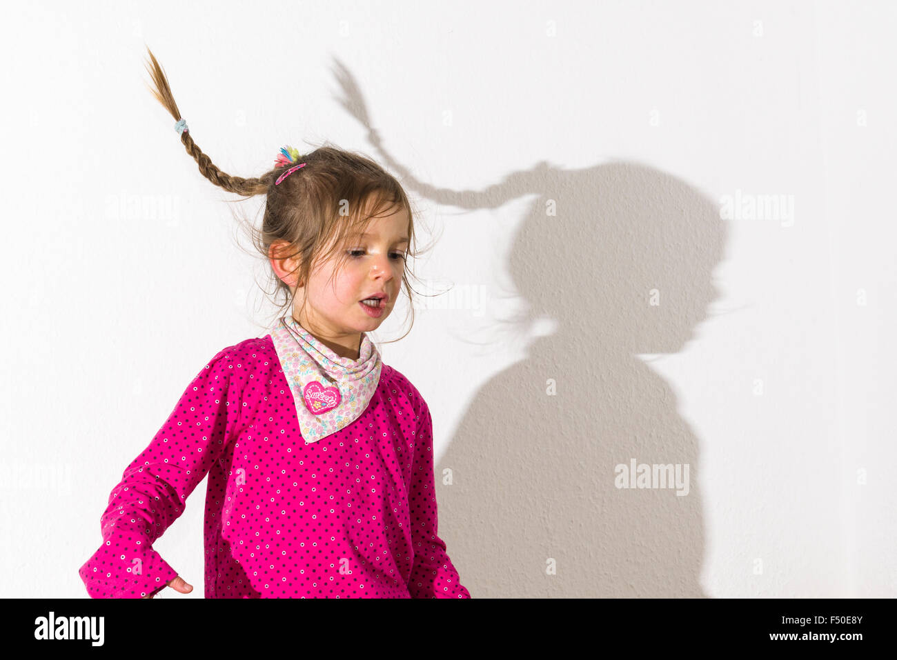 A blond three year old girl, wearing a pink shirt, is dancing in front of a white wall, leaving her shadow behind her Stock Photo