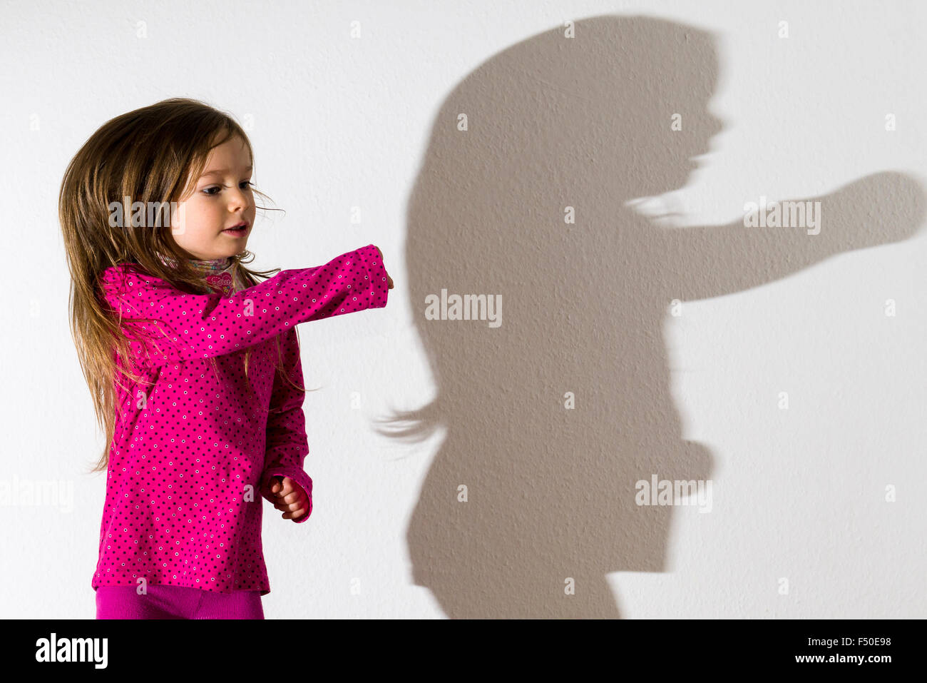A blond three year old girl, wearing a pink shirt, is dancing with open hair in front of a white wall, leaving her shadow behind Stock Photo