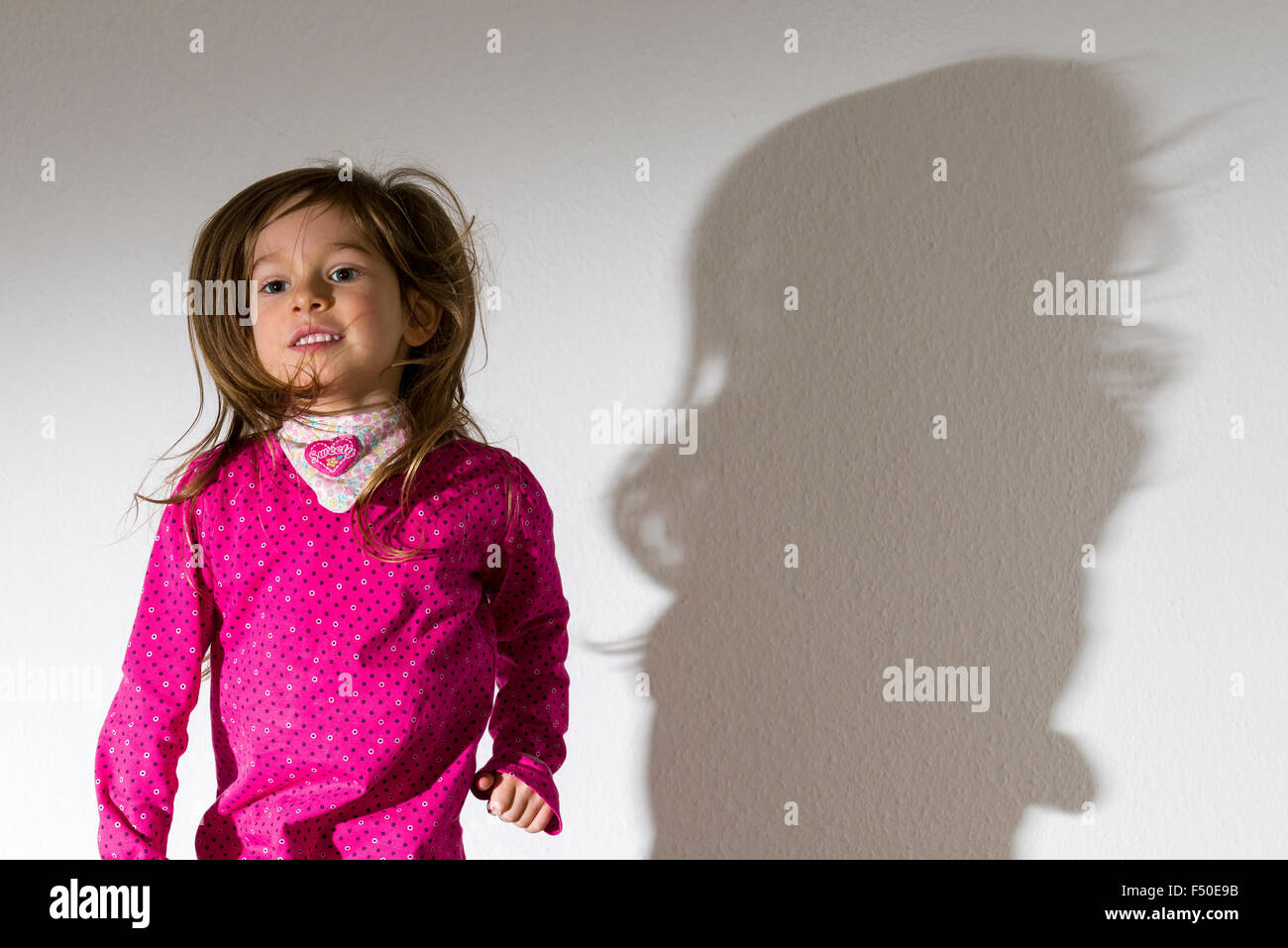 A blond three year old girl, wearing a pink shirt, is dancing with open hair in front of a white wall, leaving her shadow behind Stock Photo