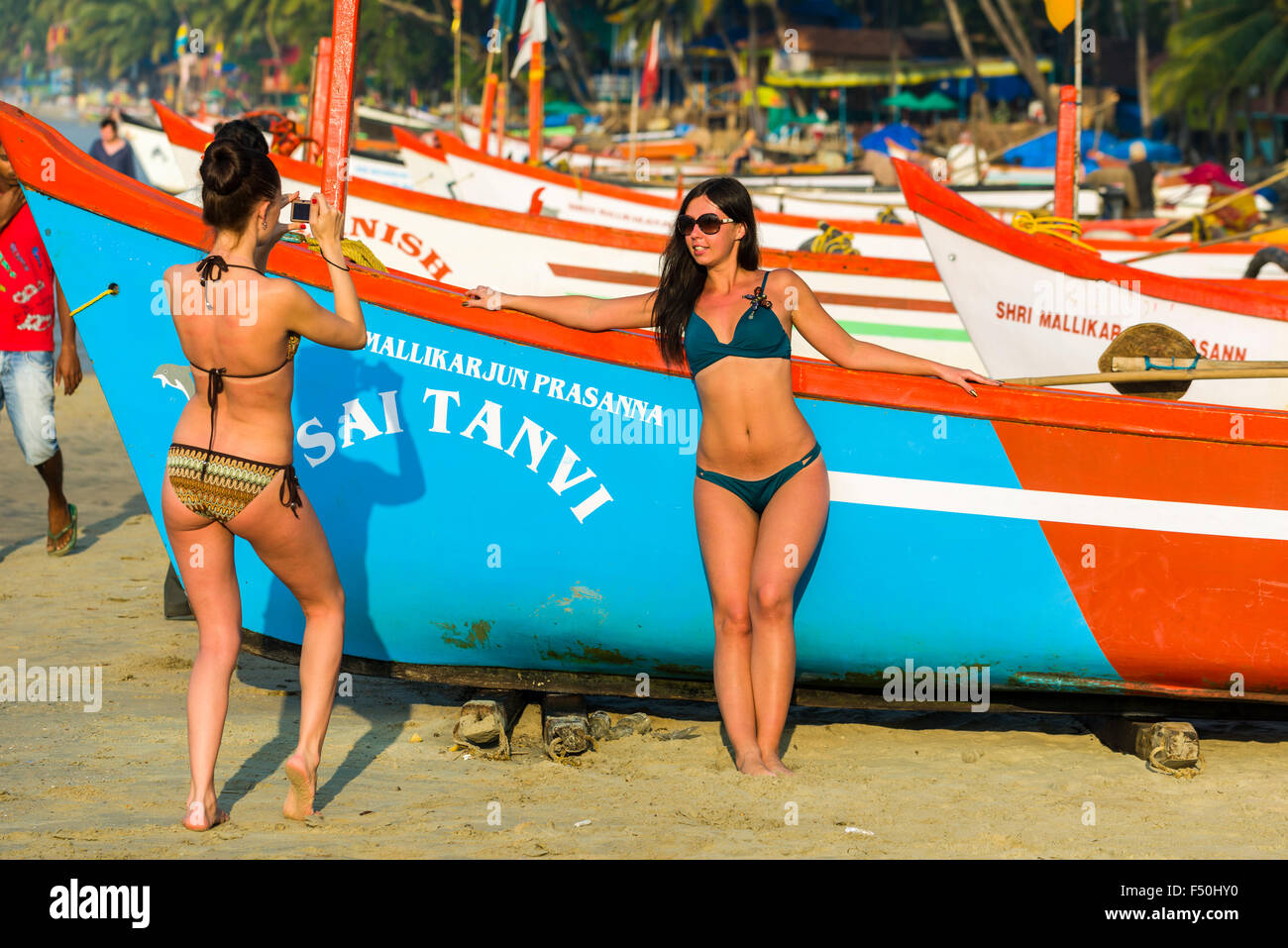 Two young women, wearing bikinis, are taking photographs of each other in front of a fishing boat at Palolem Beach Stock Photo