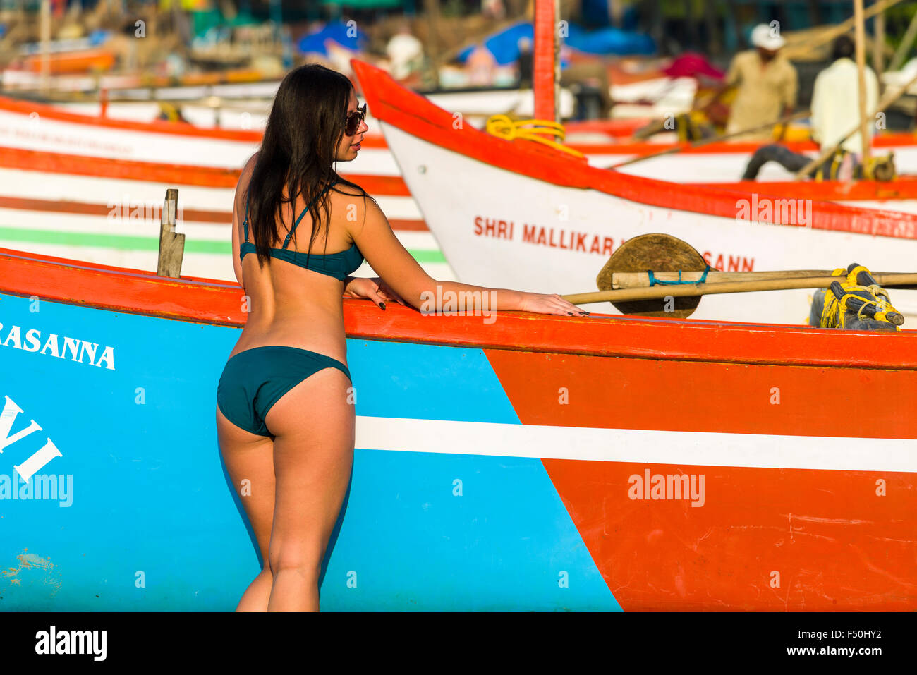 A young woman, wearing bikini, is posing for photographs in front of a fishing boat at Palolem Beach Stock Photo